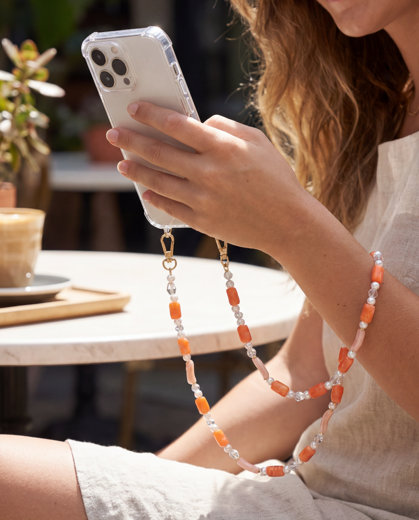 Woman holds her phone crossbody using the coral beaded Loreto strap while seated at an outdoor cafe.
