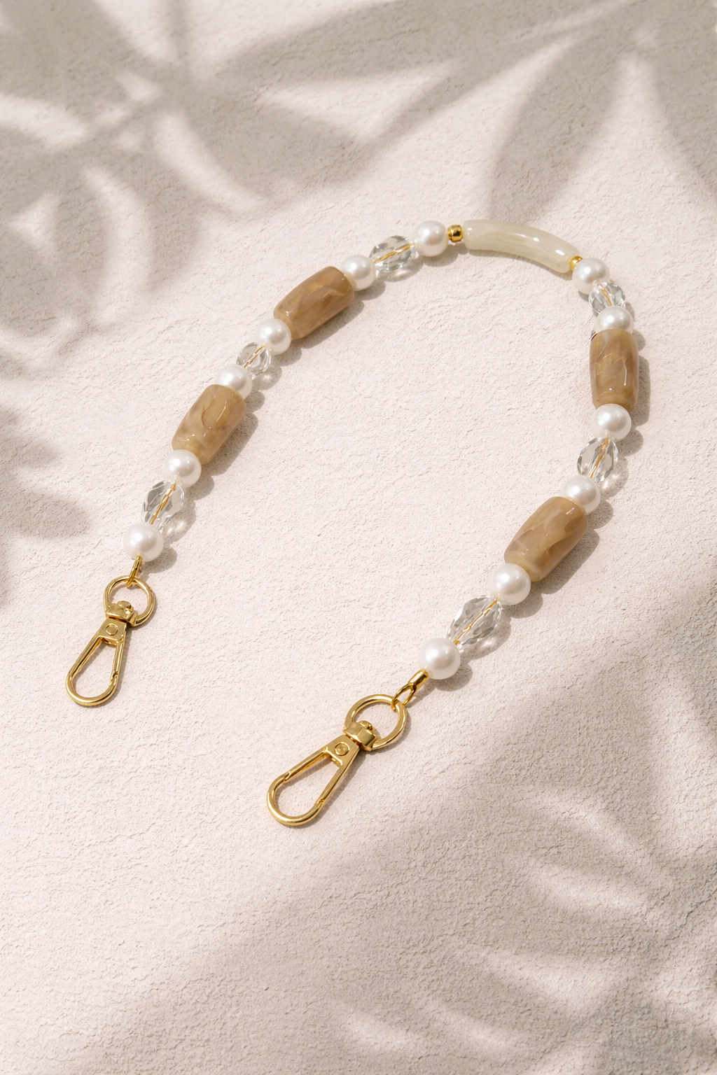 Tan beaded Loreto short phone strap styled flat on a textured surface with soft leaf-shadow lighting.