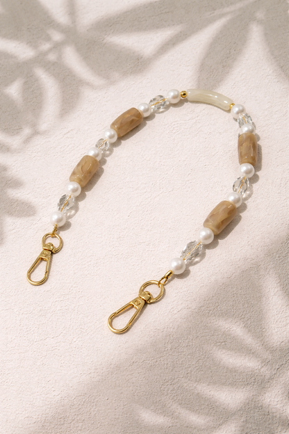 Tan beaded Loreto short phone strap styled flat on a textured surface with soft leaf-shadow lighting.