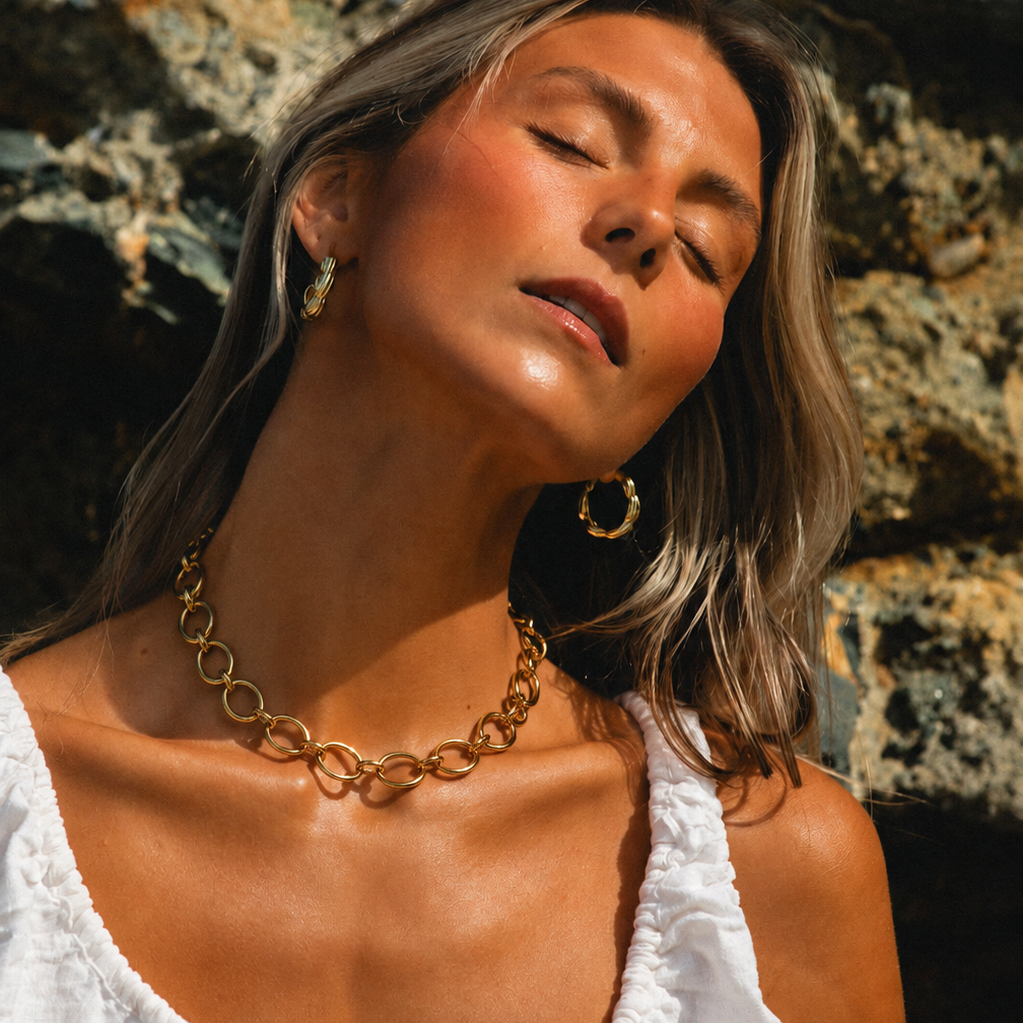 Model wears the gold Loved and Lost NECKLACE chunky oval-link chain close to the neckline, in warm sunlight on rocks.