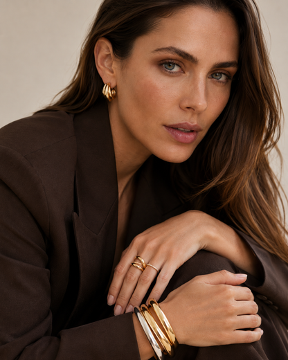 Model in a brown blazer wears the gold Lustro EARRINGS ribbed half-hoop stud, paired with stacked rings and bangles.