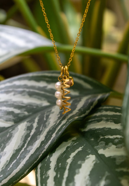 The Mama CHARM worn with a small gem and pearl accent, the gold MAMA pendant resting on a striped green houseplant leaf.