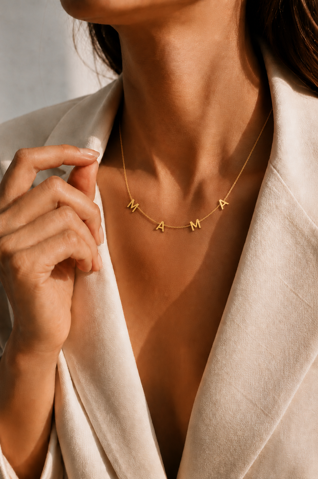 The Mama NECKLACE in gold worn at the neckline, individual letters spelling MAMA beneath a cream blazer collar.