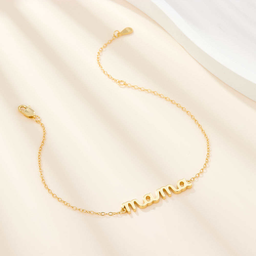 Mama Small BRACELET gold chain bracelet with cursive mama charm, draped on cream fabric near a white tray.