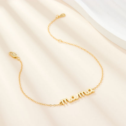 Mama Small BRACELET gold chain bracelet with cursive mama charm, draped on cream fabric near a white tray.