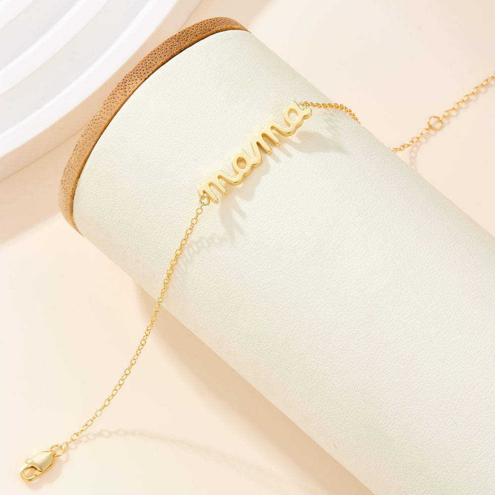 Mama Small BRACELET gold chain bracelet with cursive mama charm, styled on a leather-topped display stand.