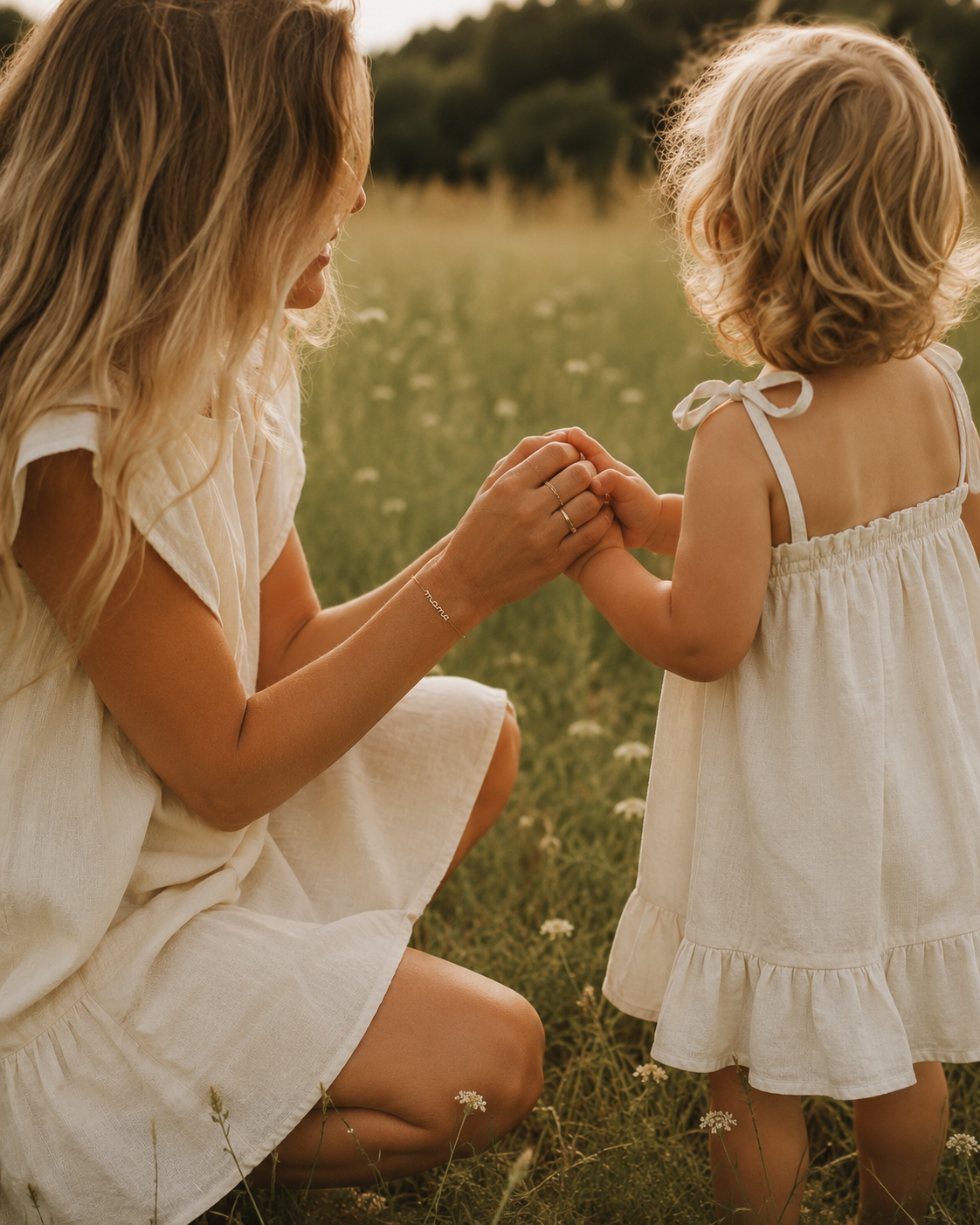 Mother and daughter in matching white dresses kneel together in a wildflower field holding hands, the mother wearing the Folie à Trois Mama Small BRACELET.