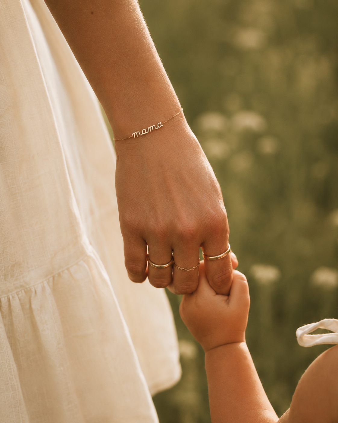 Close-up of a mother's wrist wearing the Folie à Trois Mama Small BRACELET and stacked gold rings, her fingers holding her baby's hand.