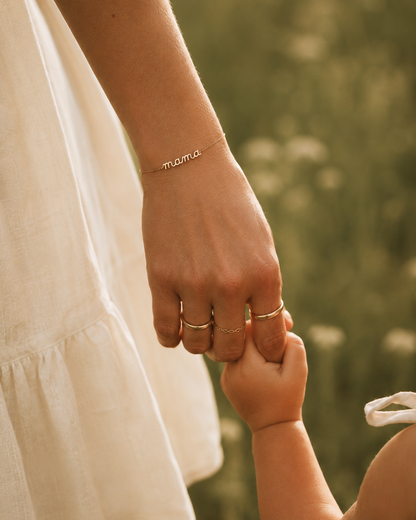 Close-up of a mother's wrist wearing the Folie à Trois Mama Small BRACELET and stacked gold rings, her fingers holding her baby's hand.