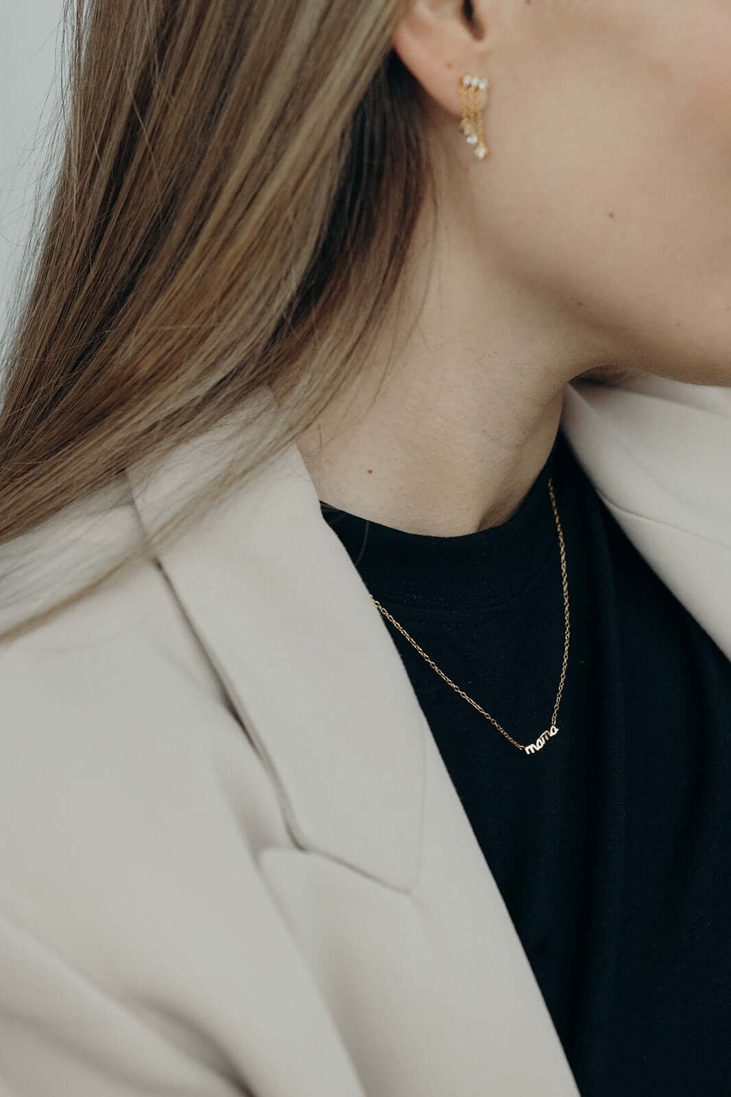 The Mama Small NECKLACE in gold worn small at the neckline under a black top and beige blazer, script mama pendant.