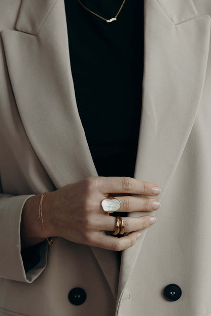 The Mama Small NECKLACE in gold worn faintly in the background of a lifestyle shot featuring stacked rings and a blazer.