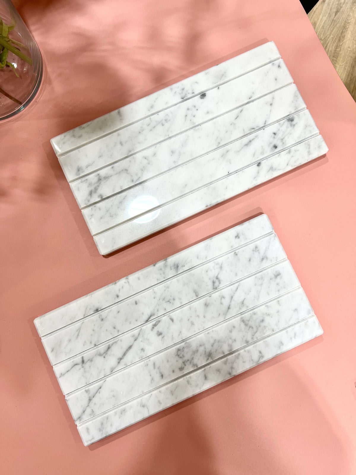 Two white marble trays with ridged slots arranged flat on a dusty pink tabletop beside a small plant.