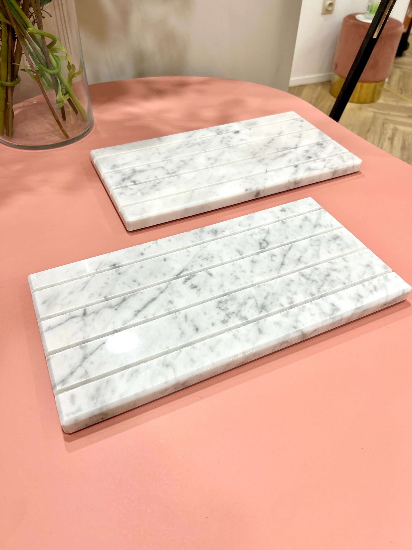 Two white marble trays with ridged slots angled on a pink table near a small potted plant.