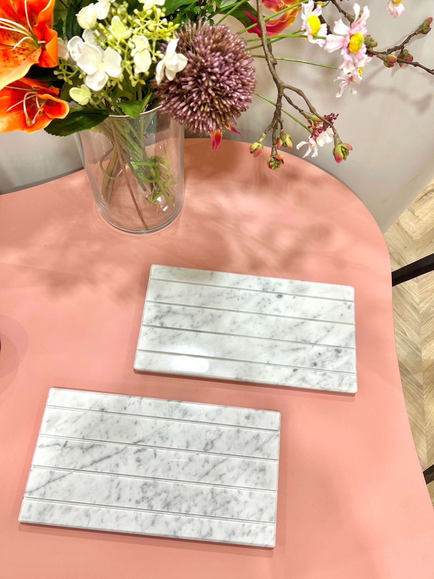 Two white marble jewelry trays on a pink table with a vase of colorful flowers in the background.