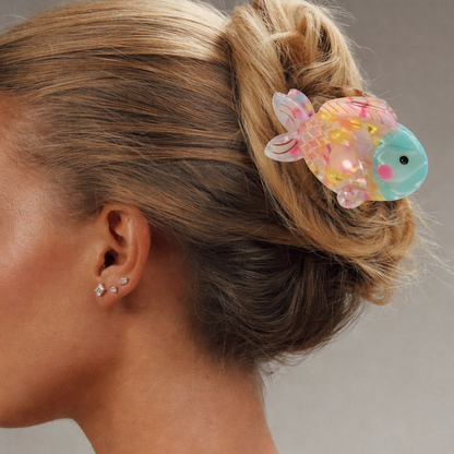 Marlin HAIR CLIP fish-shaped claw clip worn in a low bun on blonde hair, pink, yellow, turquoise colorway, with star stud earrings visible.