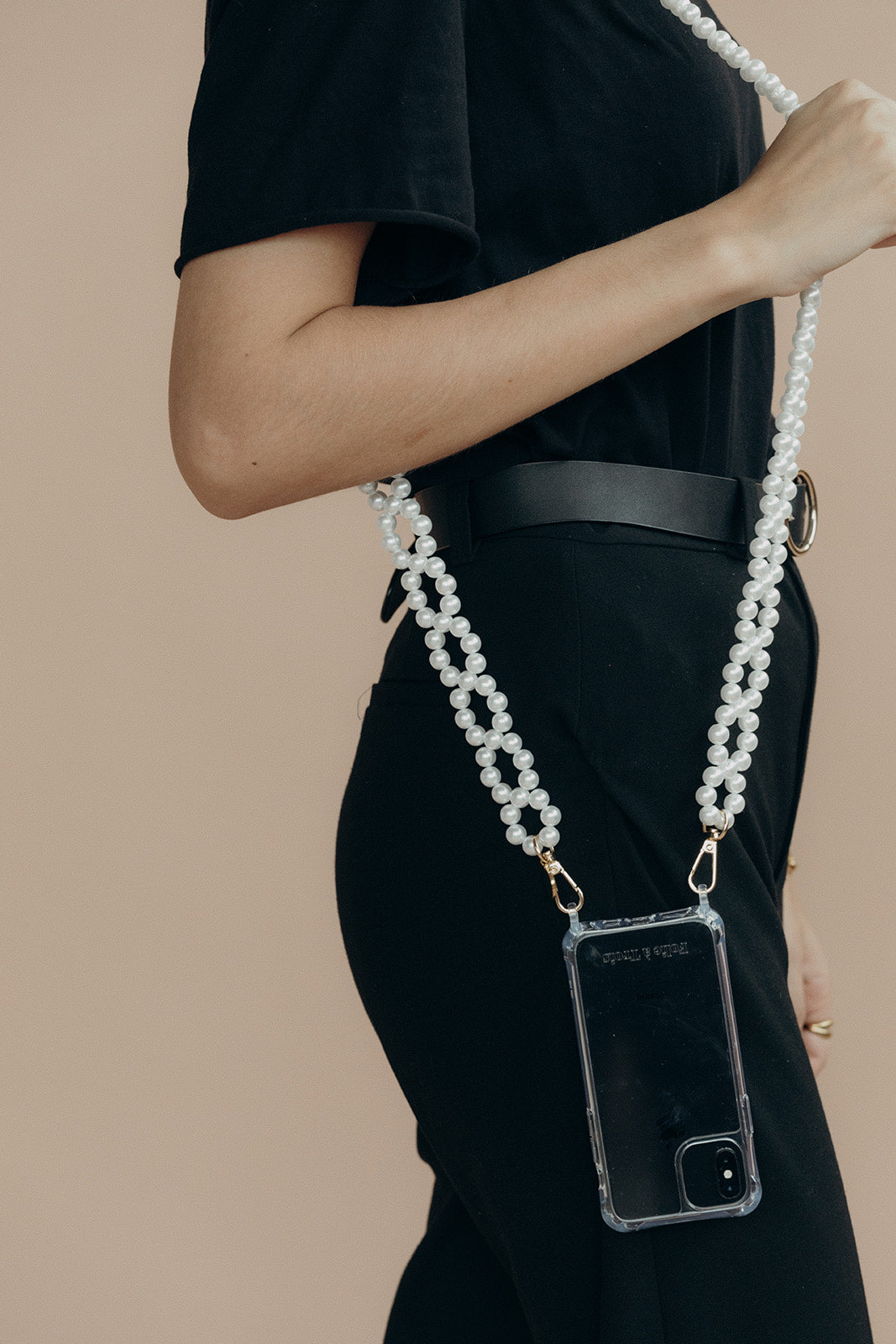 Model in black holding a phone in a clear case on the Folie à Trois Marnie Pearl wholesale phone chain, shown crossbody at the hip.