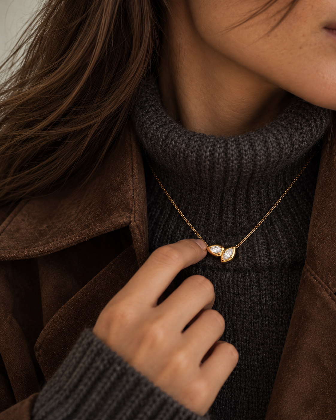 Woman touching her gold Marquise NECKLACE, layered over a grey ribbed turtleneck and brown suede jacket — Folie à Trois