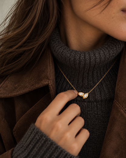 Woman touching her gold Marquise NECKLACE, layered over a grey ribbed turtleneck and brown suede jacket — Folie à Trois