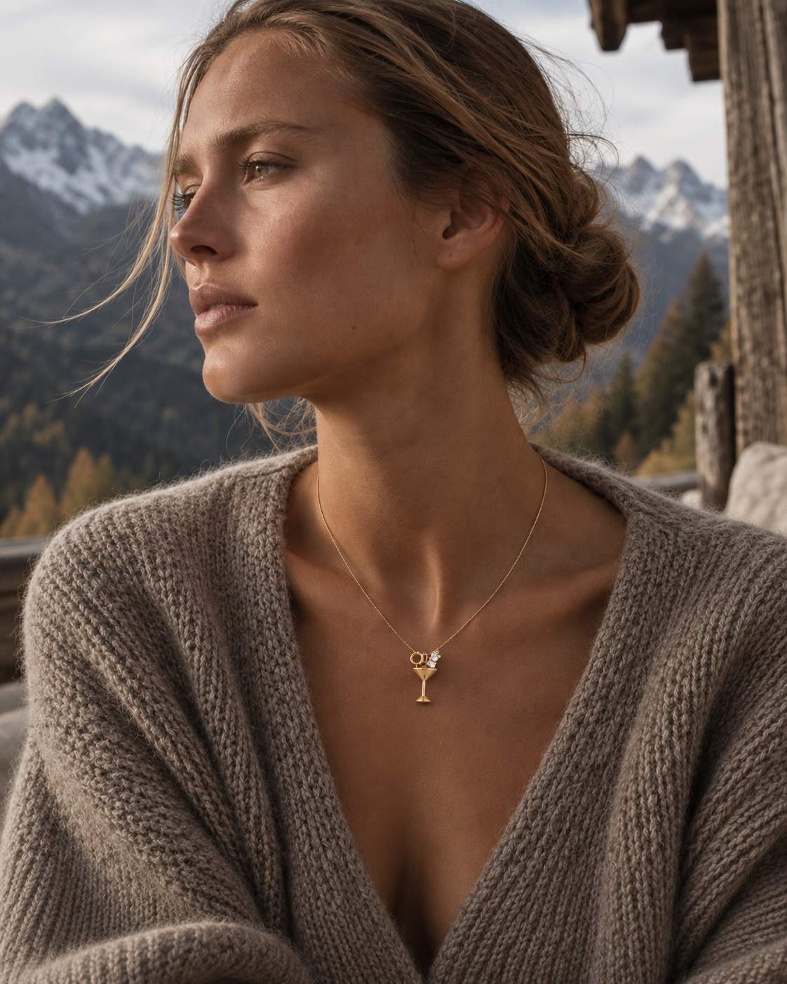 Model in a chunky taupe knit cardigan wearing the gold Martini Glass CHARM on a fine chain, with snowy mountains behind her.