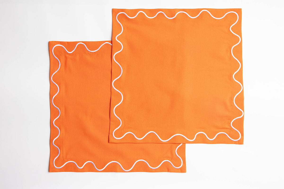 Two folded orange Maude napkins with white wave-trim edging, stacked flat on a white background.