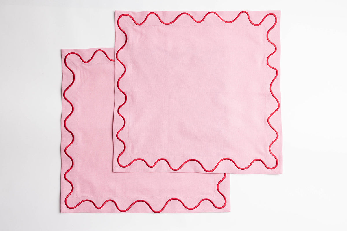 Two folded pink Maude napkins with a red scalloped wave-trim edge, stacked neatly on a white background.