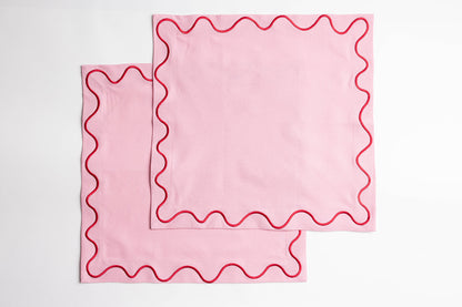 Two folded pink Maude napkins with a red scalloped wave-trim edge, stacked neatly on a white background.