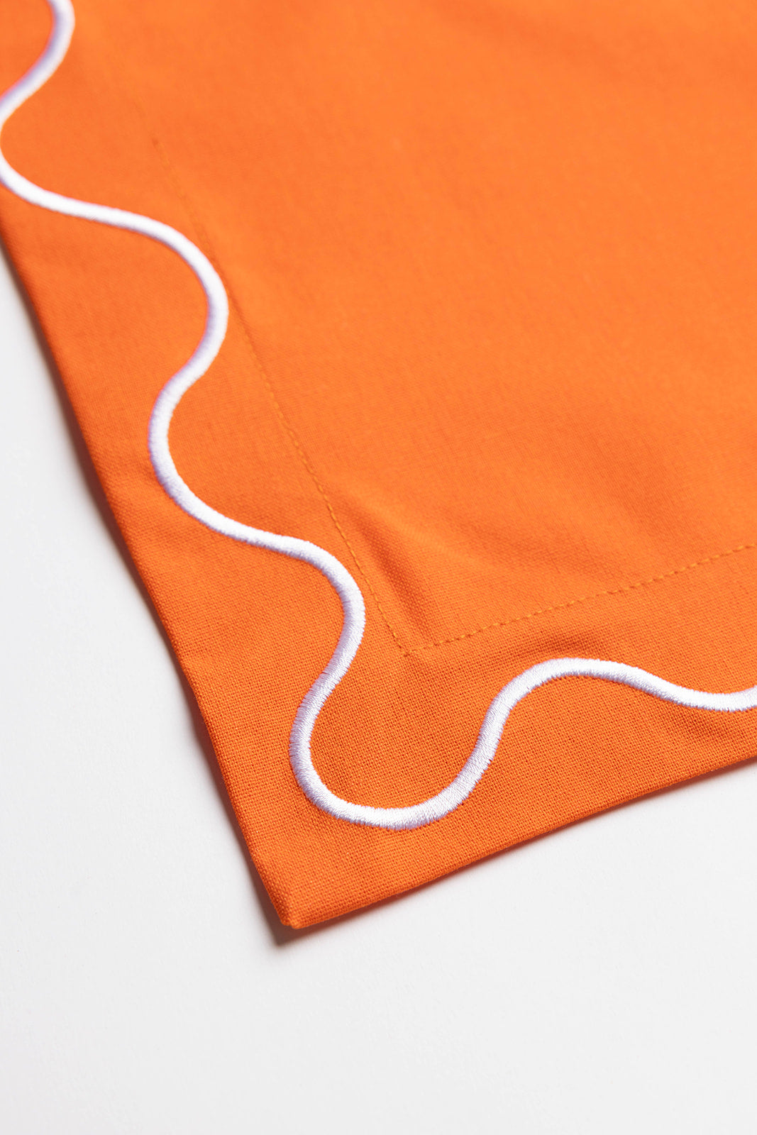 Close-up corner of an orange Maude napkin showing the white scalloped wave-trim stitching detail.