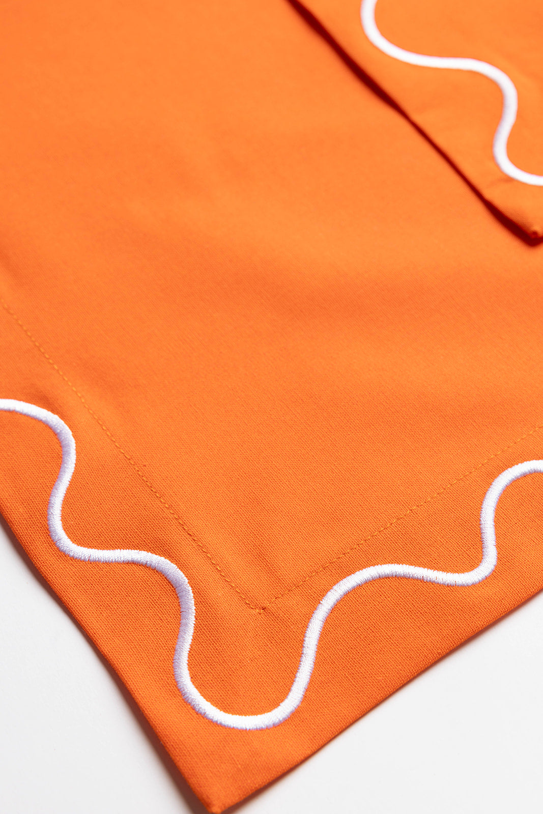 Close-up folded corner of an orange Maude napkin, highlighting the white scalloped wave-trim edge.