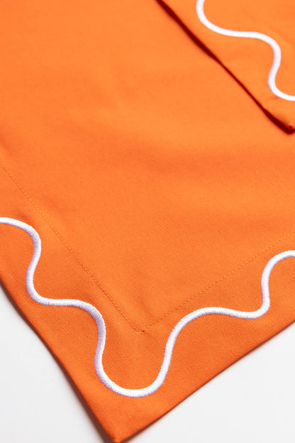 Close-up folded corner of an orange Maude napkin, highlighting the white scalloped wave-trim edge.