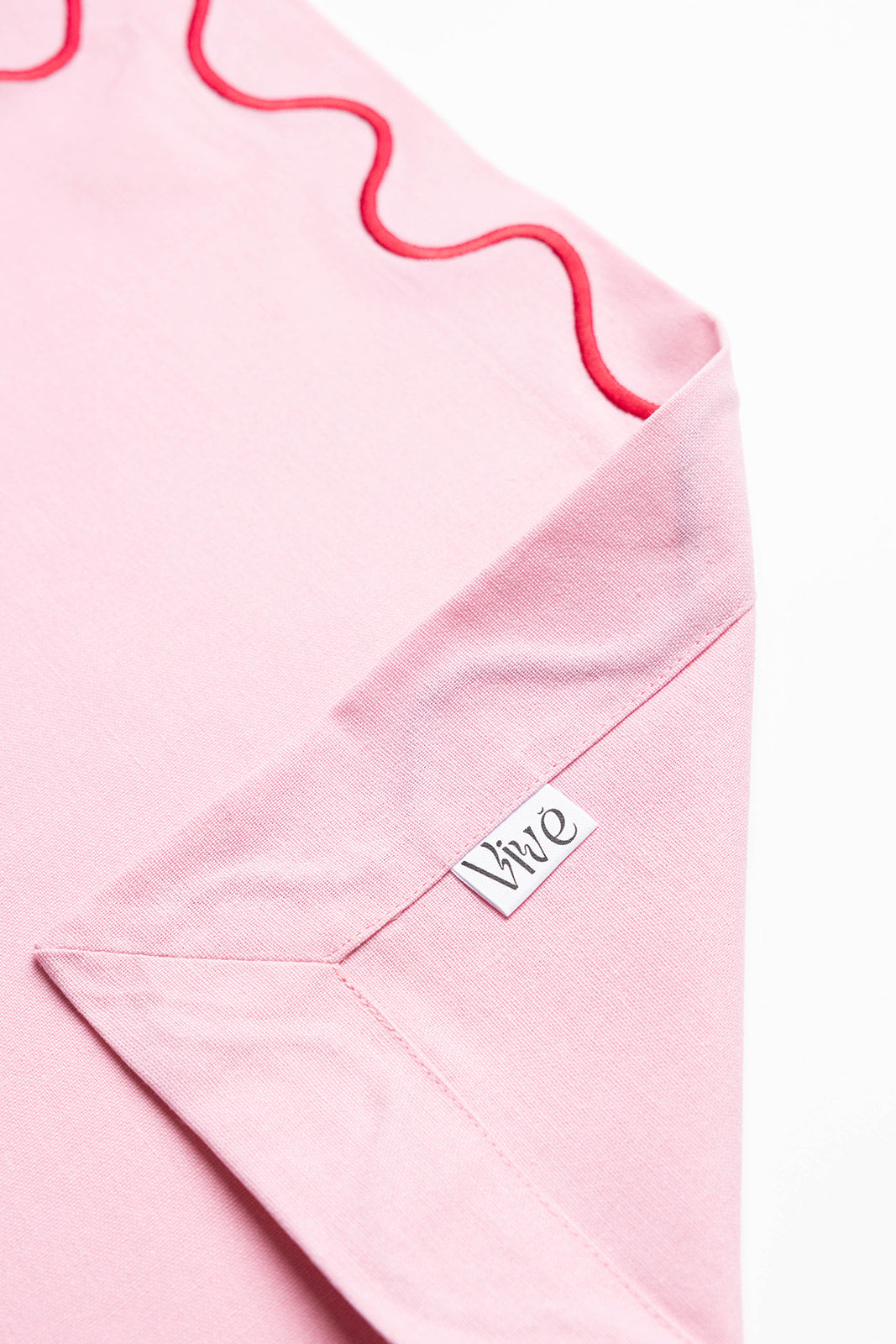 Close-up of a pink Maude napkin corner with red wave trim and the folded Vive brand label tag visible.