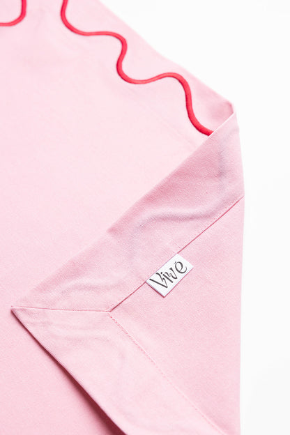 Close-up of a pink Maude napkin corner with red wave trim and the folded Vive brand label tag visible.