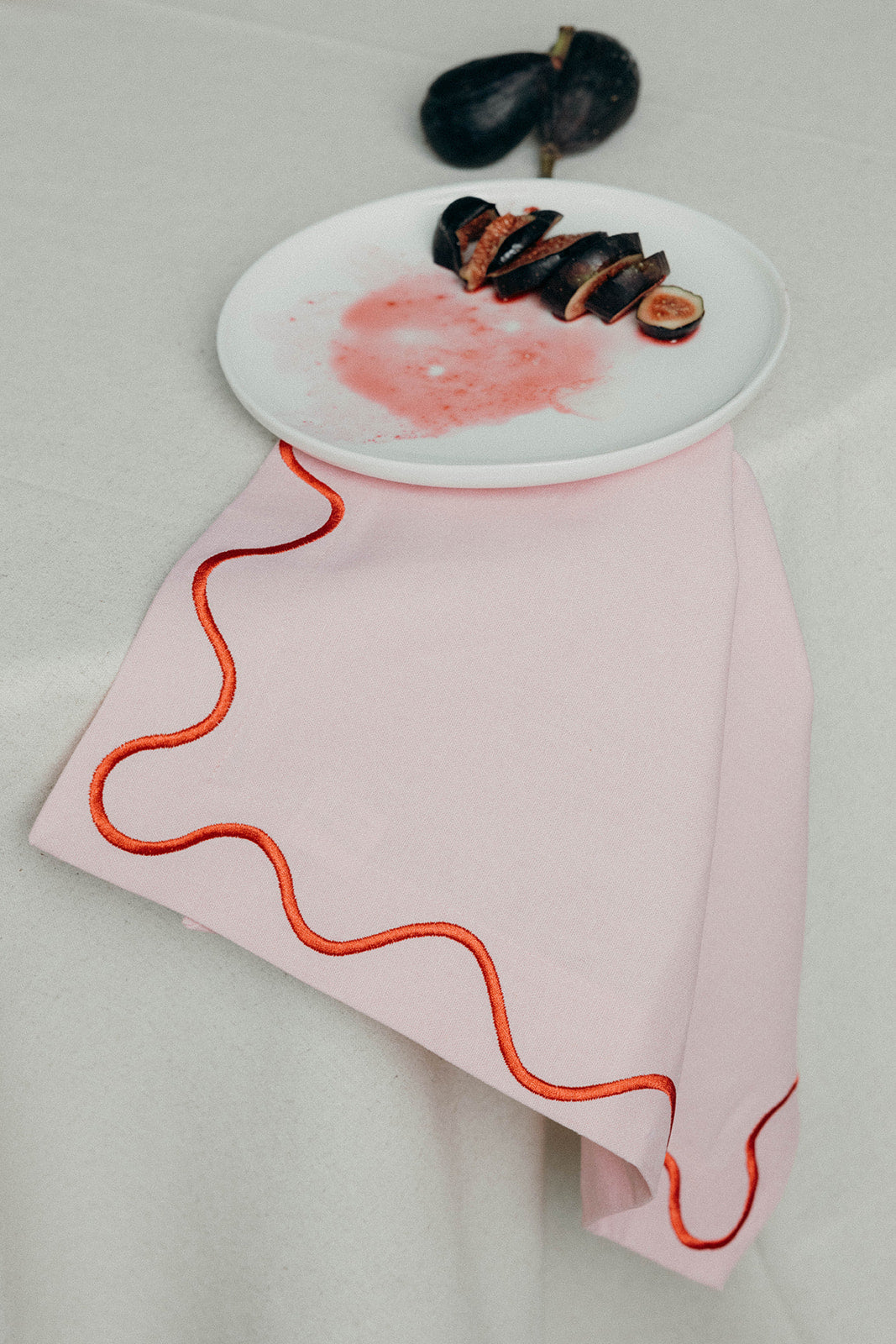 Pink Maude napkin with an orange wave-trim border draped beneath a white plate of sliced fresh figs.