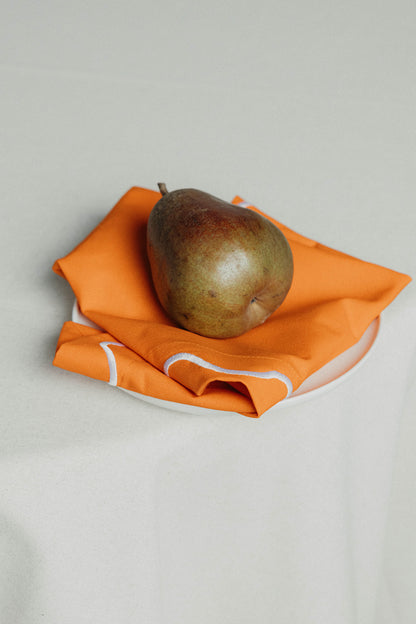A pear resting on a folded orange Maude napkin with white wave-trim edging, set on a white plate.