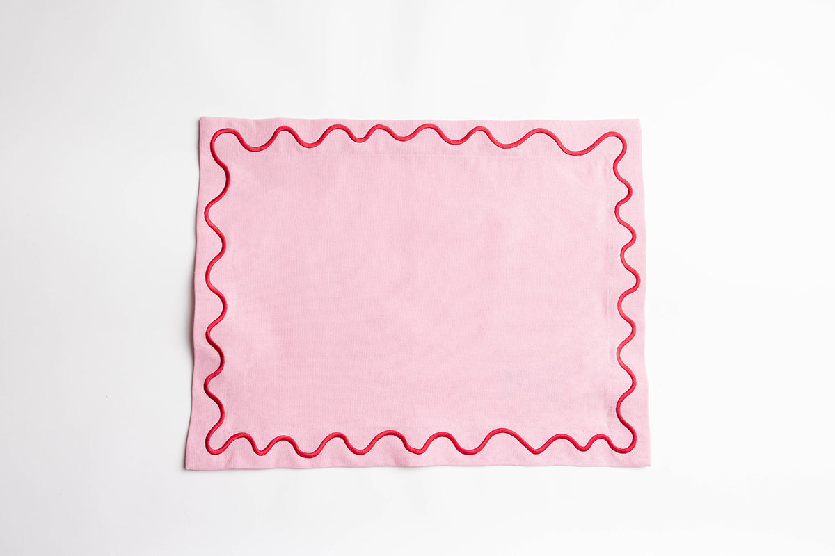 Pink Maude placemat with a red scalloped wave-trim border, laid flat on a white surface to show the embroidered edge detail.