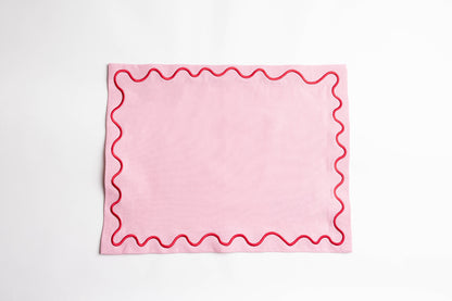 Pink Maude placemat with a red scalloped wave-trim border, laid flat on a white surface to show the embroidered edge detail.