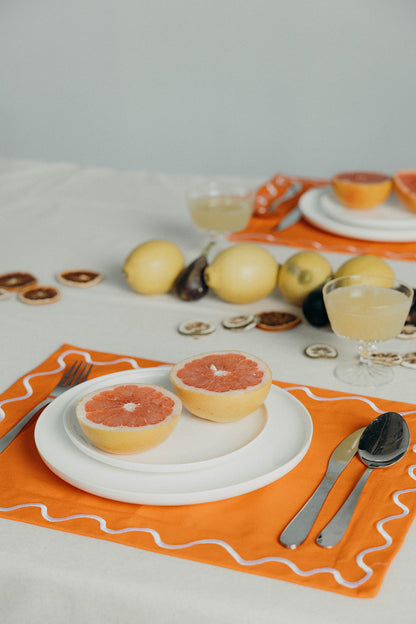 Orange Maude wave-trim placemat styled with a white plate of halved grapefruit, cutlery and a citrus cocktail glass.