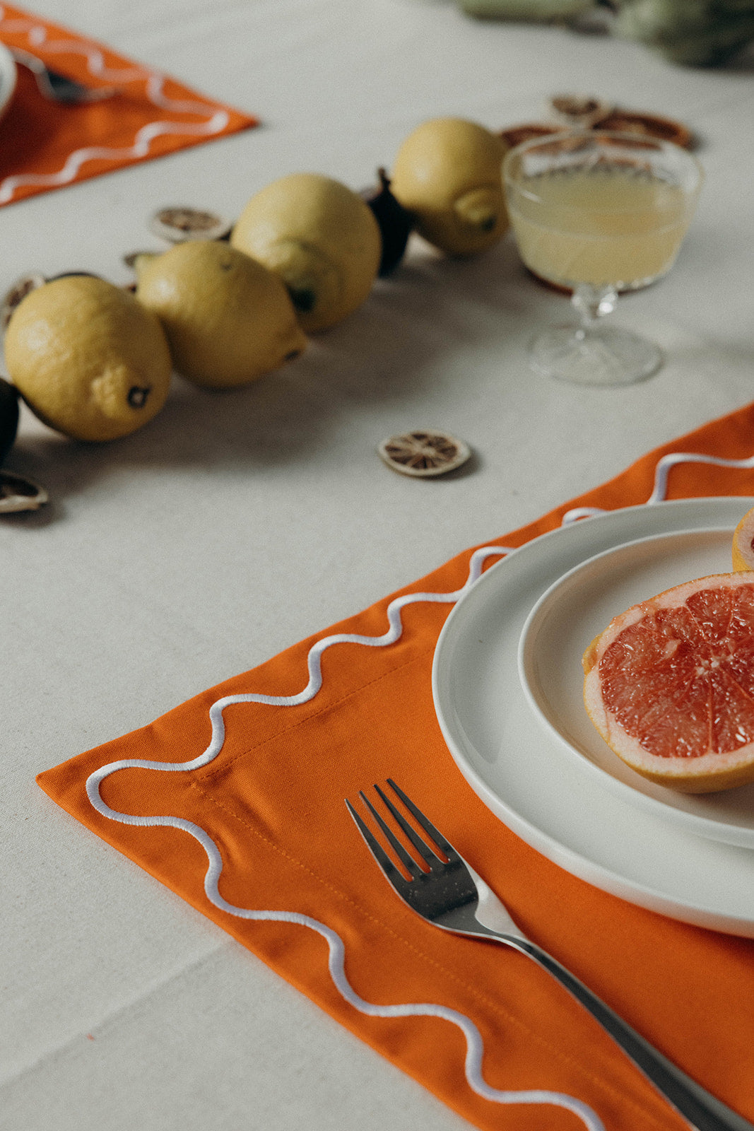 Close-up of an orange Maude wave-trim placemat holding a grapefruit-topped plate, with lemons and a cocktail nearby.