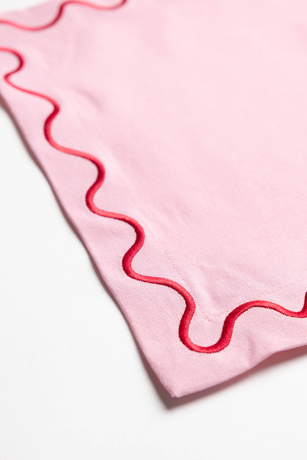 Close-up corner of a pink Maude placemat showing its red scalloped wave-trim stitching in fine detail.