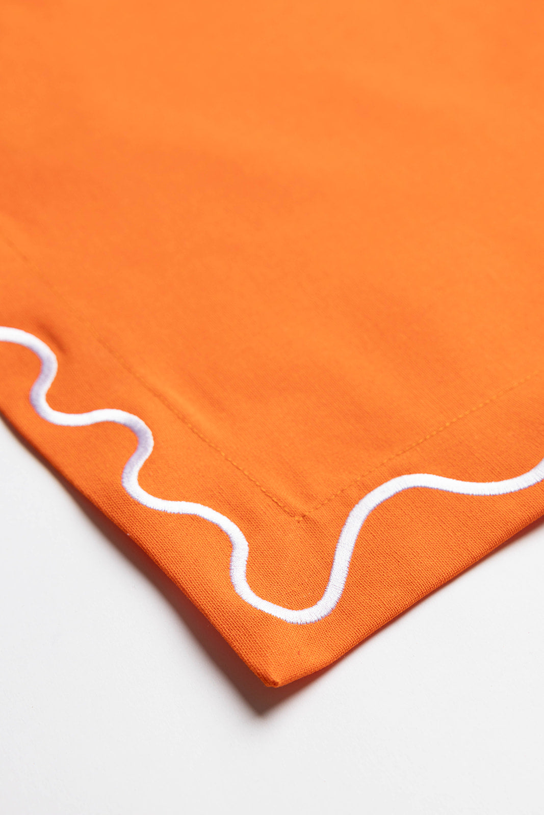 Close-up corner of an orange Maude placemat highlighting the white scalloped wave-trim embroidery.