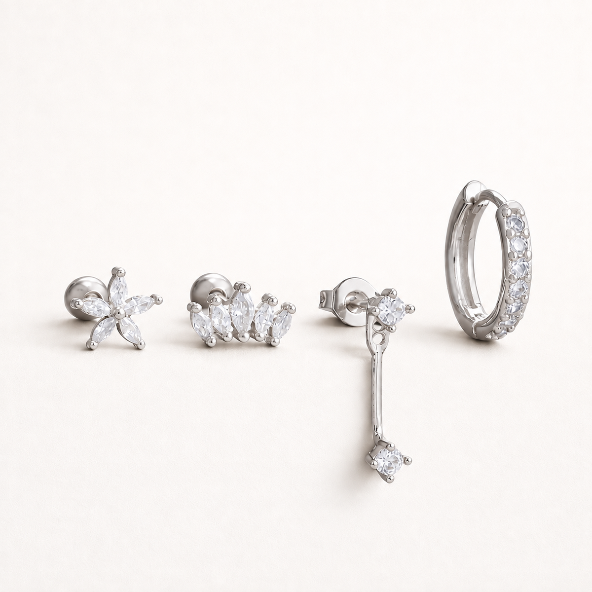 Four silver Melanie SET pieces laid flat: a marquise flower stud, a marquise cluster stud, a chain-drop stud and a pavé huggie hoop.