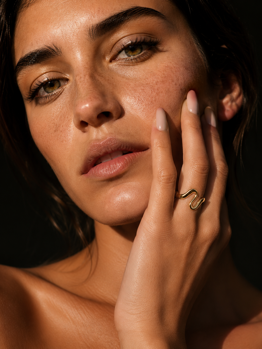 Model wears the gold Menton RING, its slender S-curve visible on her finger as she touches her cheek in dramatic side lighting.