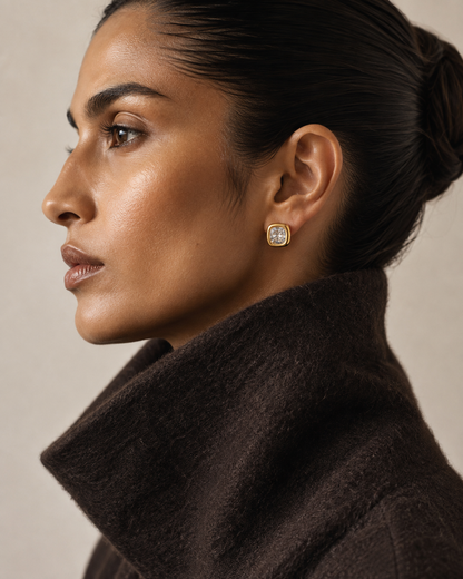 Profile view of a model in a brown coat wearing the gold Meridian EARRINGS, a cushion-cut zirconia bezel stud.