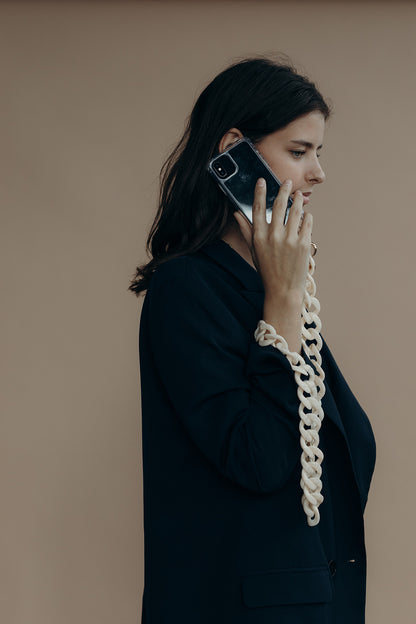 A woman on the phone wearing a cream twisted-link phone chain that drapes down the sleeve of her dark blazer.