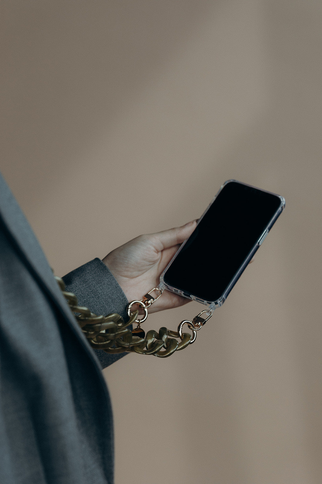 A hand holding a phone by its olive green curb-link chain, against a blurred beige background.