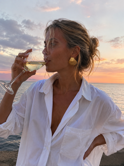 Model wears the Folie à Trois Midday Sun EARRINGS, a gold scallop-shell stud, sipping wine at golden-hour by the sea.