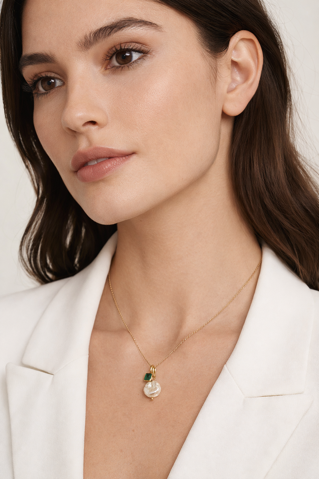 The Minimal NECKLACE worn close to the collarbone with a pearl charm cluster and a small square emerald charm, white blazer visible.