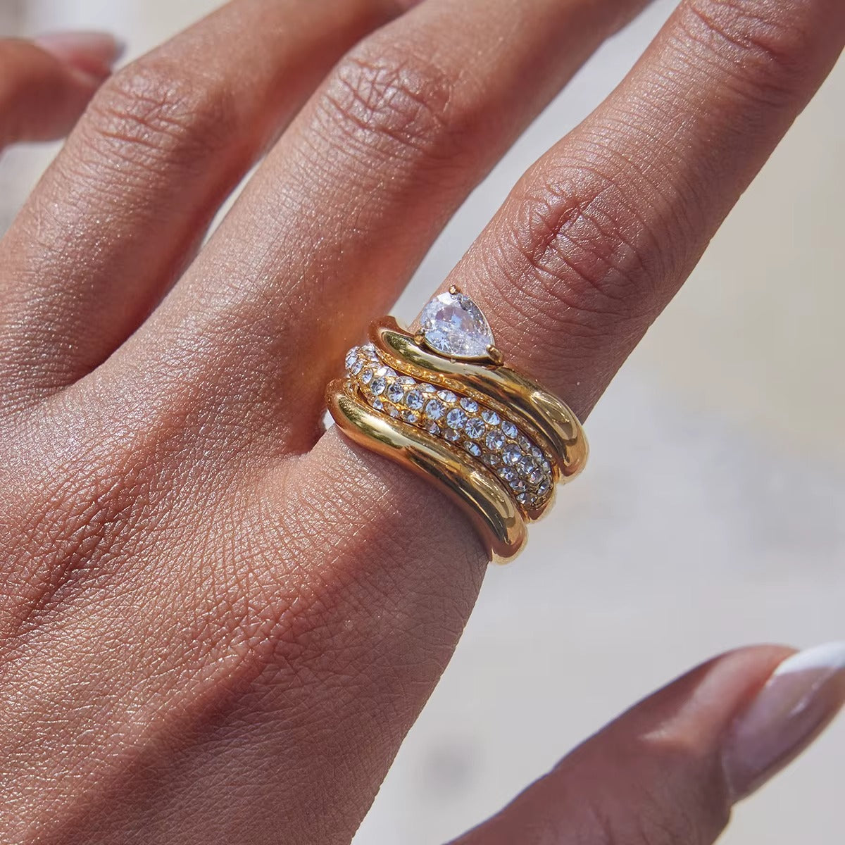 Close-up of a hand stacked with three gold Misbehave rings — a pear-stone band, a pavé band and a plain wavy band — on one finger.
