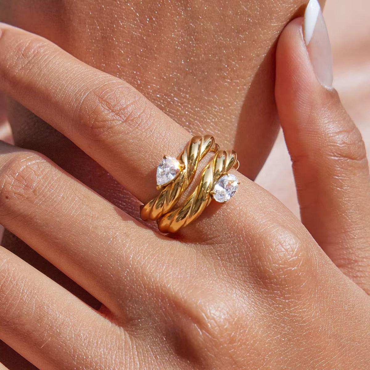 Close-up of a hand near the neck wearing two twisted gold Misbehave rings, each set with a clear pear-shaped stone, stacked together.