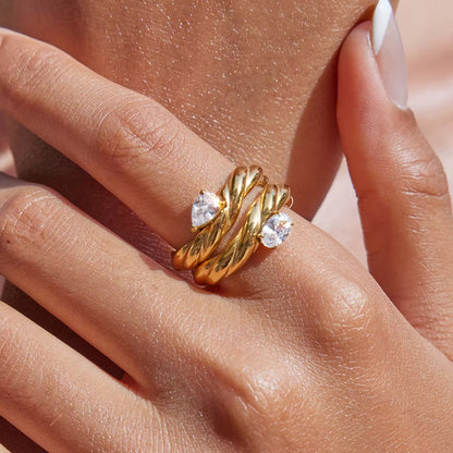 Close-up of a hand near the neck wearing two twisted gold Misbehave rings, each set with a clear pear-shaped stone, stacked together.