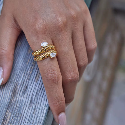 Hand resting on weathered wood wearing two twisted gold Misbehave rings with clear pear-shaped stones stacked on one finger.